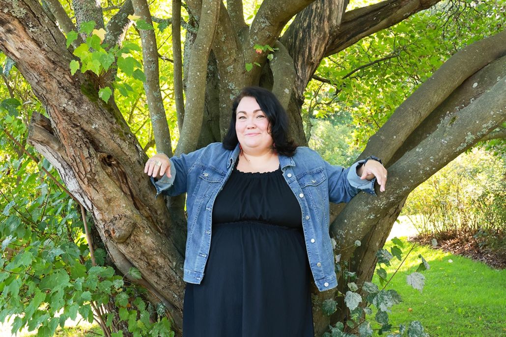 Kuvassa hymyilevä nainen nojaamassa puunrunkoon