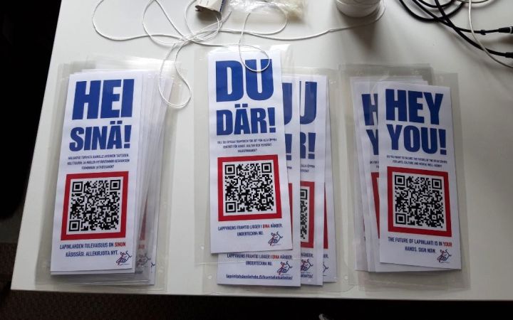Laminoidut Hei sinä! -julisteet QR-koodilla valkoisella pöydällä