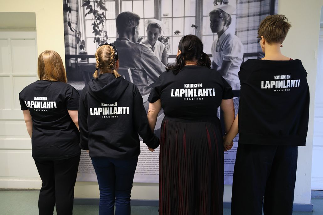 Kuvassa neljä ihmistä katselee Mental Museum-näyttelyä yllään paidat, joiden selkämyksissä lukee Kaikkien Lapinlahti.