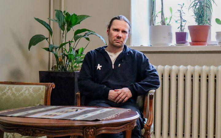 Kuvassa miesoletettu istuu puisen pöydän ääressä. Pöydällä on pöytäliina, ja taustalla näkyy viherkasveja.