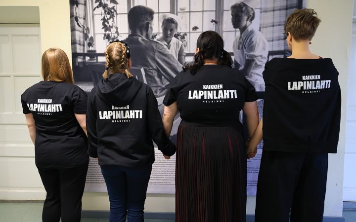 Kuvassa neljä ihmistä katselee Mental Museum-näyttelyä yllään paidat, joiden selkämyksissä lukee Kaikkien Lapinlahti.