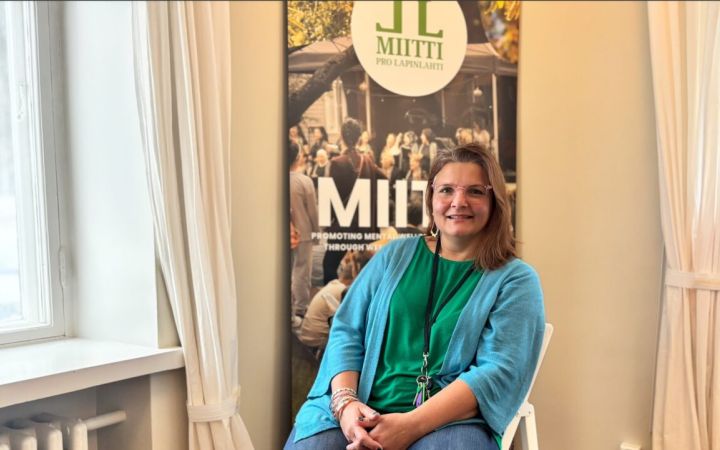 Nainen istuu MIITTI-julisteen edessä tuolissa ja hymyilee.