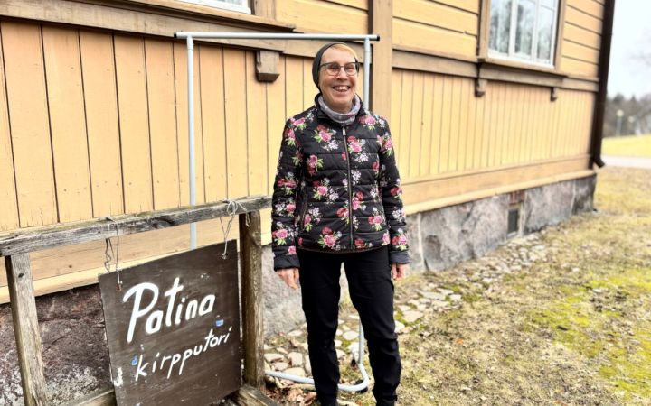 Nainen hymyilee ja seisoo ulkona puisen rakennuksen edessä kukikkaassa takissa.