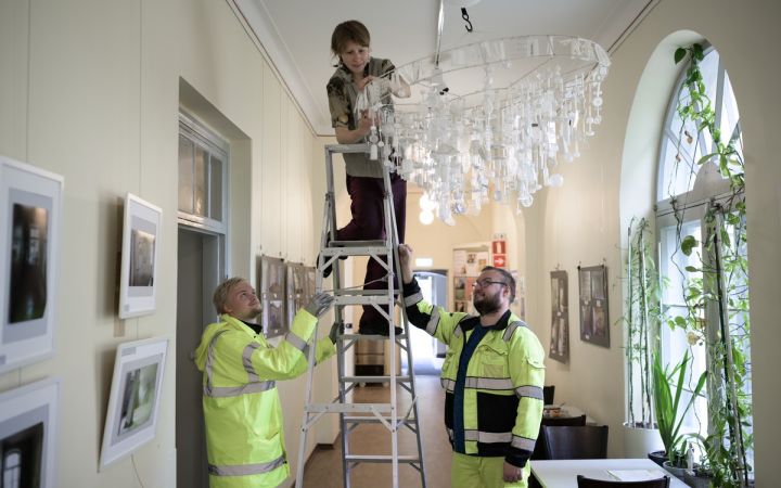 Nainen asentaa valaisinteosta tikkailla kahden henkilön avustamana.