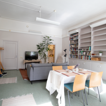 Kuvassa on Valo-tila, etualalla on pöytä ja tuoleja, taeempana sohva ja televisio. Oikealla on kirjahylly.