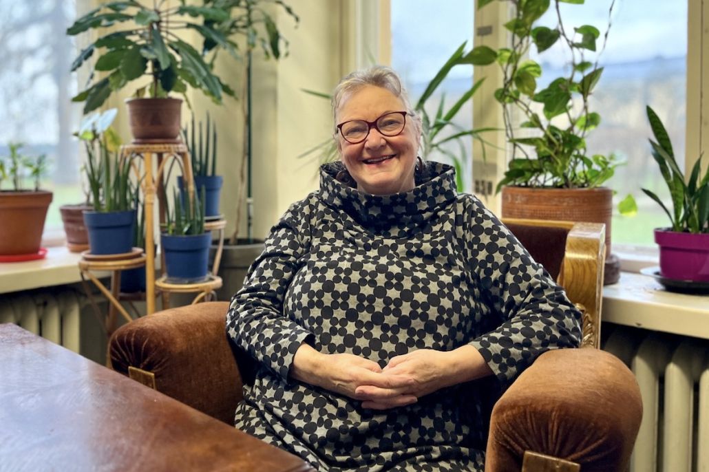 Nainen hymyilee tuolissa viherkasvien ja ikkunan äärellä.