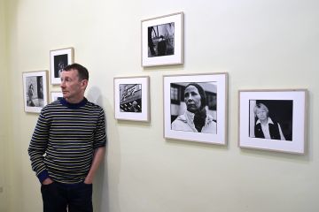 Raitapaitainen mies nojaa seinään, seinällä mustavalkoisia valokuvatauluja Aki Kaurismäen näyttelijöistä.