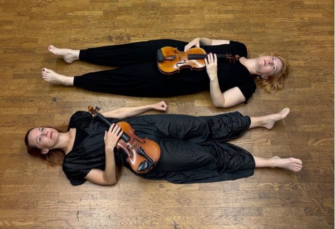 Kaksi musta-asuista naista lepää lattialla viulut sylissä.
