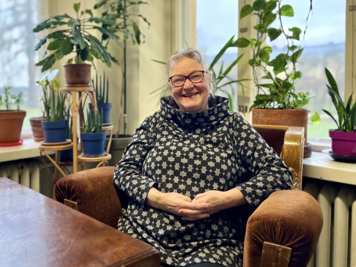 Nainen hymyilee tuolissa viherkasvien ja ikkunan äärellä.