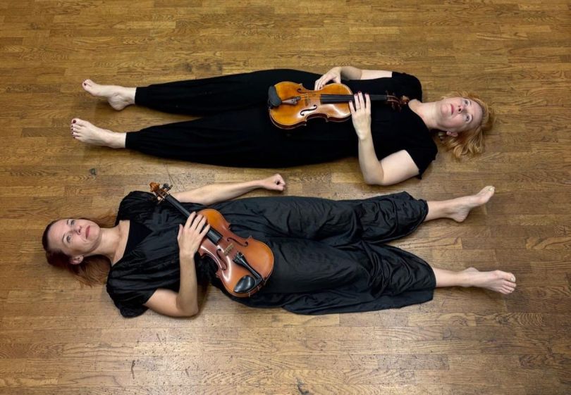 Kaksi musta-asuista naista lepää lattialla viulut sylissä.