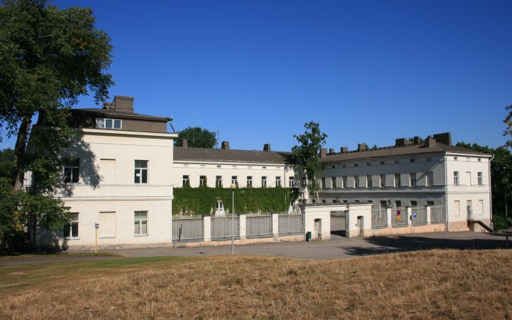 Lapinlahden sairaalarakennus kuvattuna etuviistosta. Etualalla kuivaa nurmikkoa ja taustalla sininen taivas.