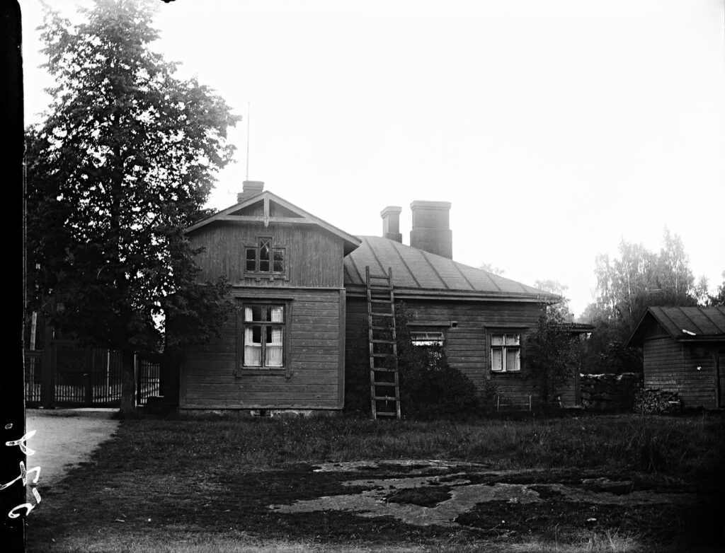 Mustavalkoisen kuvan keskellä puinen portinvartijan rakennus. Kuvattu sairaalan puolelta. Vasemmalla sairaala-alueen portti.