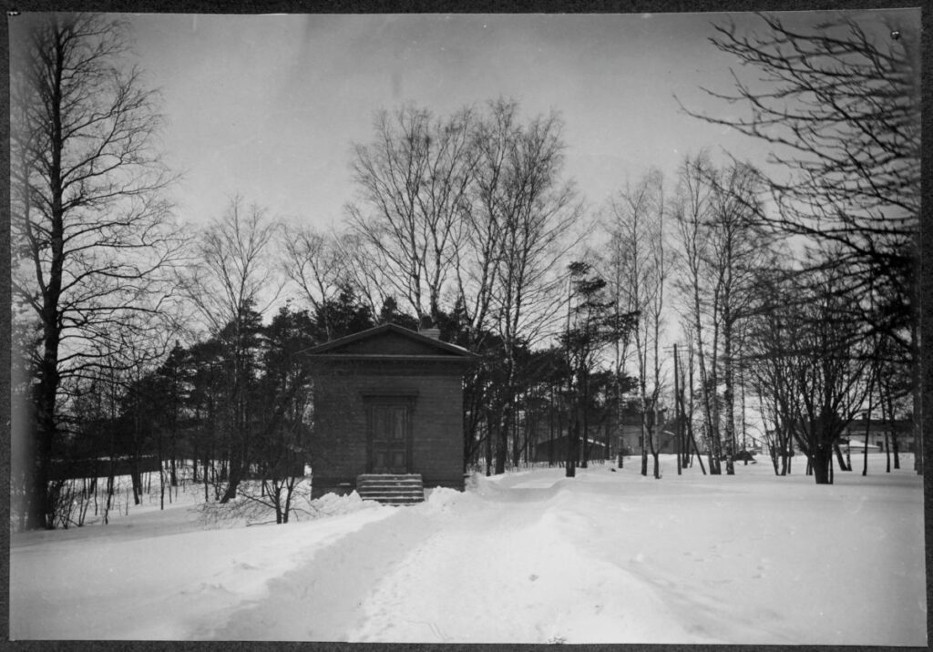 Aika pimeä mustavalkoinen kuva. Kuvassa näkyy puita ja siunauskappeli. Maassa on lunta.