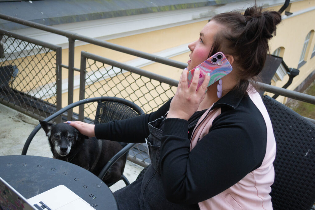 Nainen istuu parvekkeella ja puhuu puhelimessa, samalla kun rapsuttaa viereisellä tuolilla olevaa koiraa.