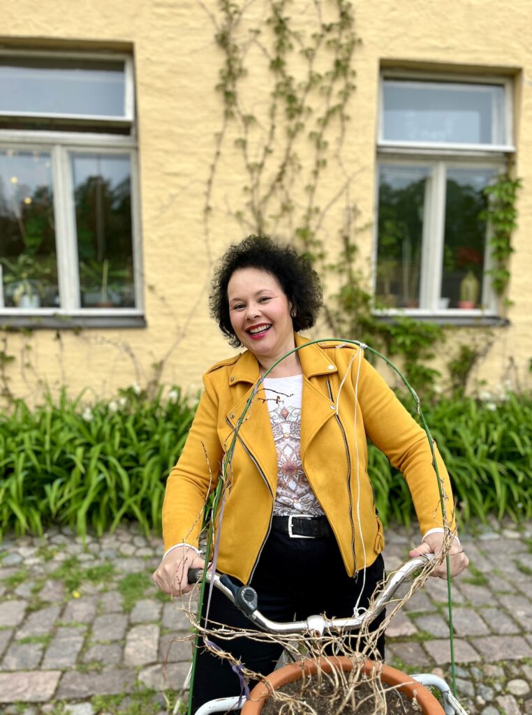 Keltatakkinen nainen poseeraa vanhan polkupyörän päällä keltaisen rakennuksen seinää vasten.