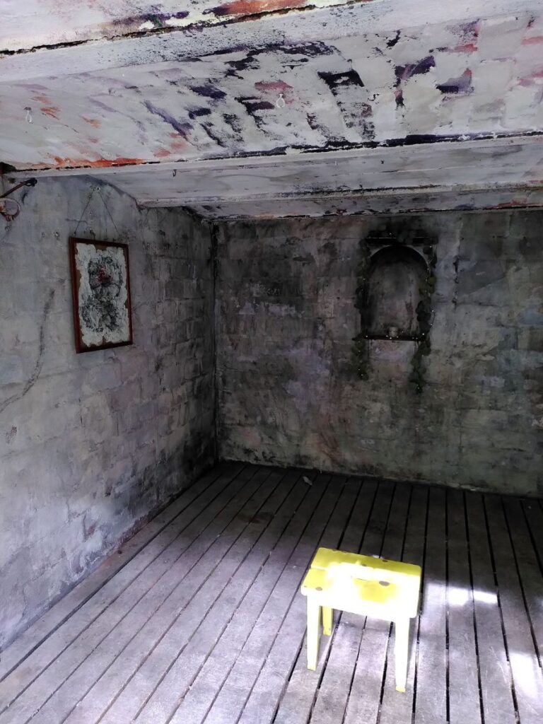 Kuvassa on kammion eli siunauskappelin kellarin sisätila. Vasemmalla seinällä on taulu, oikealla seinällä syvennys. Keskellä lattiaa auringon valossa on keltainen jakkara.