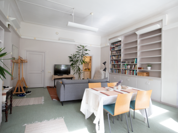 Kuvassa on Valo-tila, etualalla on pöytä ja tuoleja, taeempana sohva ja televisio. Oikealla on kirjahylly.