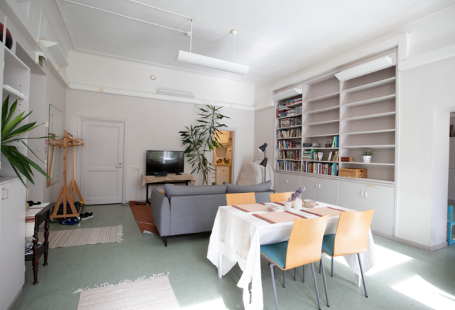 Kuvassa on Valo-tila, etualalla on pöytä ja tuoleja, taeempana sohva ja televisio. Oikealla on kirjahylly.