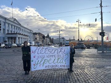 Kaksi ihmistä pitelee kylttiä kaupungintalon edustalla, taustalla nousee aurinko.