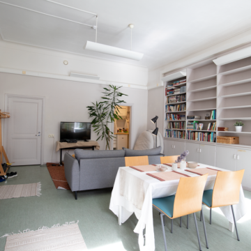 Kuvassa on Valo-tila, etualalla on pöytä ja tuoleja, taeempana sohva ja televisio. Oikealla on kirjahylly.