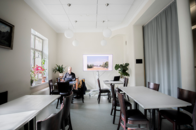 Kuvassa etualalla on pöytiä ja tuoleja. Taaempana on pöydässä istuva henkilö ja Osasto5:n televisio.