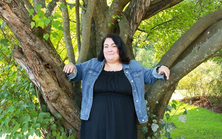 Kuvassa hymyilevä nainen nojaamassa puunrunkoon