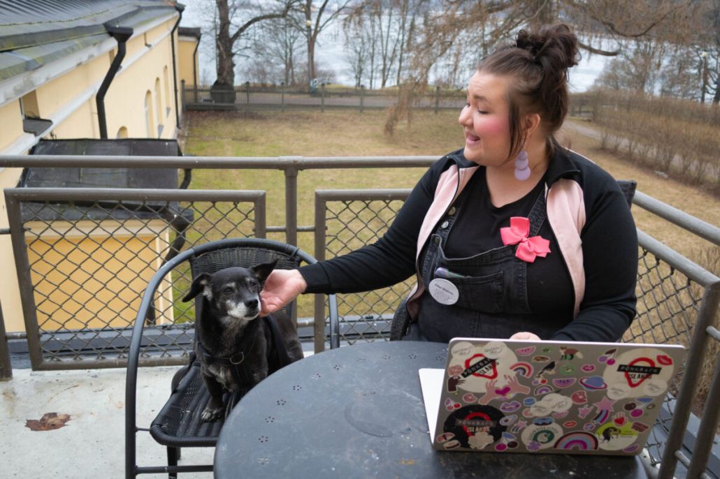 Nainen istuu parvelleella tietokoneen ääressä ja viereisesllä tuolilla on pieni musta koira.