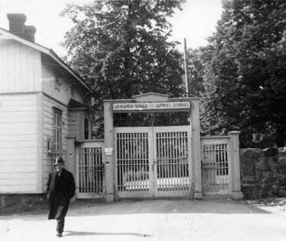 Mustavalkoisessa kuvassa mies kävelee poispäin sairaalan portilta. Portin vieressä on puinen rakennus.