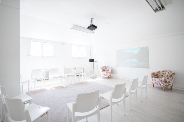 Kuvassa etu- ja taka-alalla on tuoleja. Keskellä on matto ja takaseinällä projektorin lähettämä kuva.