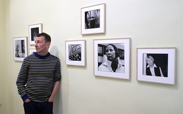 Raitapaitainen mies nojaa seinään, seinällä mustavalkoisia valokuvatauluja Aki Kaurismäen näyttelijöistä.