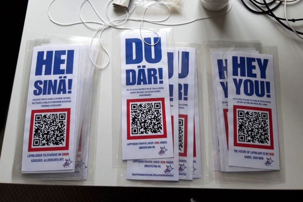 Laminoidut Hei sinä! -julisteet QR-koodilla valkoisella pöydällä