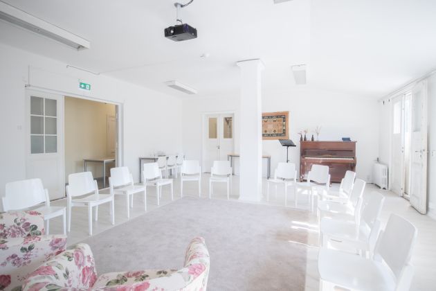 Kuvassa on tuoleja kehässä. Keskellä on matto ja taka-alalla piano.