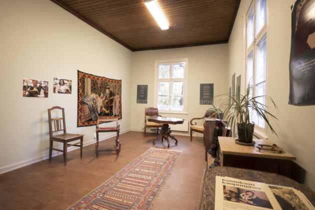 Kuvassa etualalla on matto ja seinän vierustalla huonekaluja. Seinillä on tauluja.