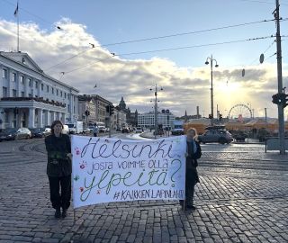 Kaksi ihmistä pitelee kylttiä kaupungintalon edustalla, taustalla nousee aurinko.