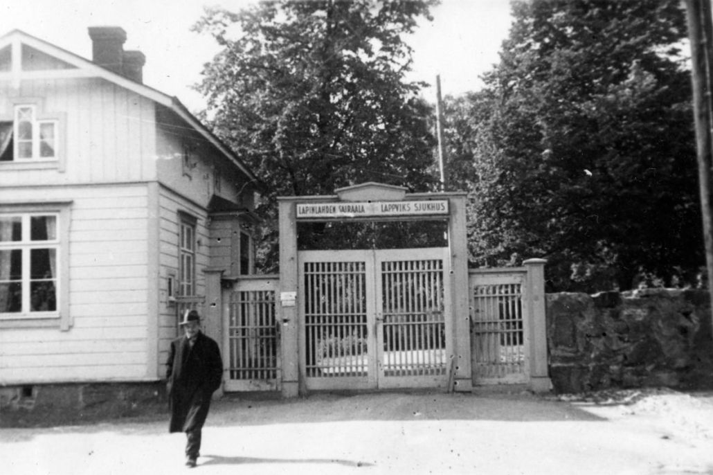 Mustavalkoisessa kuvassa mies kävelee poispäin sairaalan portilta. Portin vieressä on puinen rakennus.