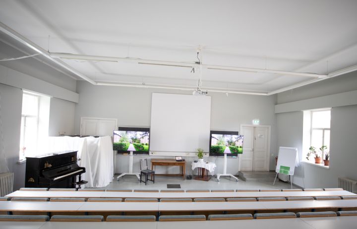 Kuvassa on auditorion valkokangas ja kaksi televisiota.