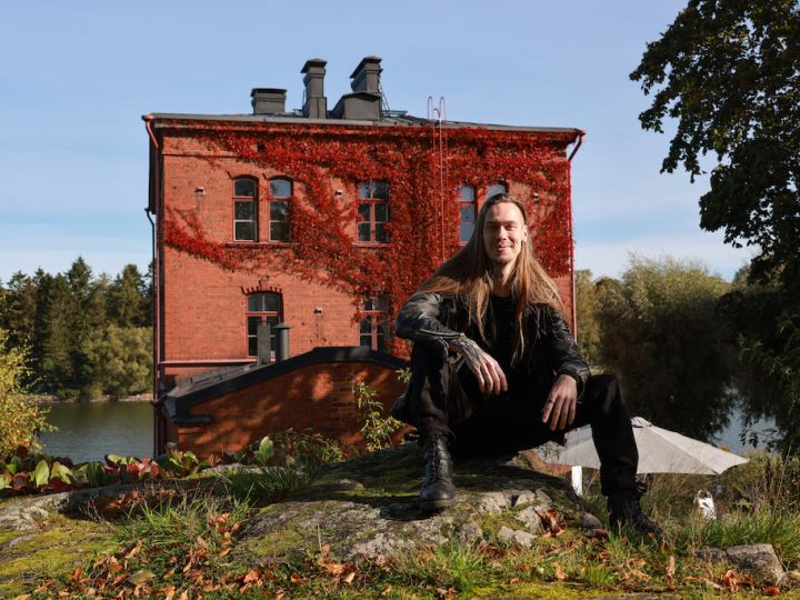 Tiilinen Venetsia-talo ja edustalla pitkähiuksinen istuva, hymyilevä mies.