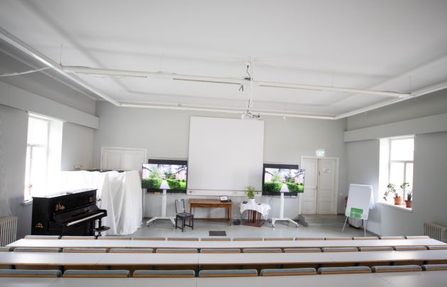 Kuvassa on auditorion valkokangas ja kaksi televisiota.