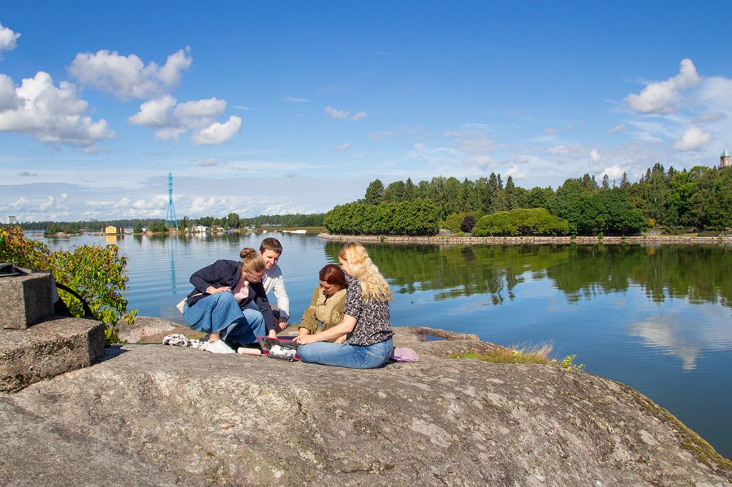 Neljä ihmistä aurinkoisilla Lapinlahden kallioilla meren äärellä keskustelemassa.