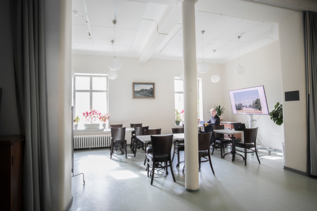 Kuvassa on Osasto5:n pöytiä ja tuoleja, etualalla on pylväitä. Taka-alalla on pöydässä istuva henkilö ja Osasto5:n televisio.