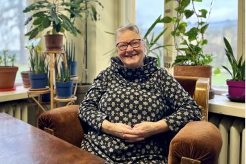 Nainen hymyilee tuolissa viherkasvien ja ikkunan äärellä.