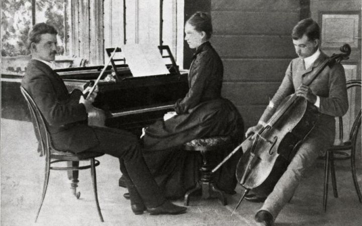 Mustavalkoinen kuva Sibeliuksen sisaruksista. Jean istuu vasemmalla sylissään viulu, Linda keskellä pianon ääressä ja Christian Sibelius oikealla sellon kanssa.