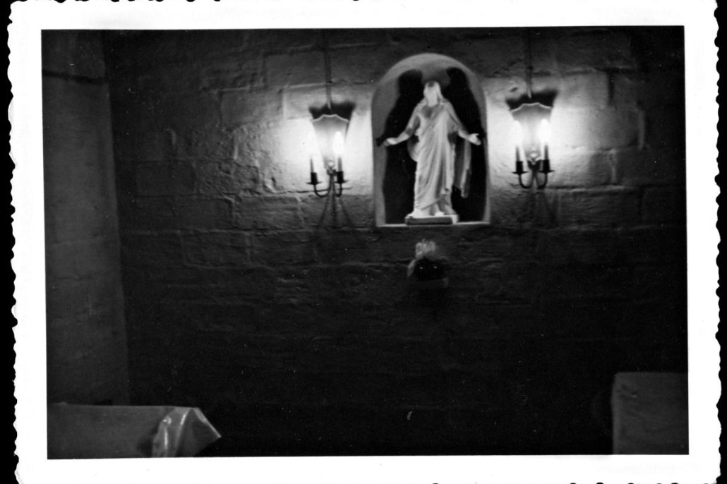 Hyvin pimeä kuva siunauskappelin sisältä. Seinällä olevassa syvennyksessä näkyy uskonnollinen patsas ja sen vierellä seinäkyntteliköt.