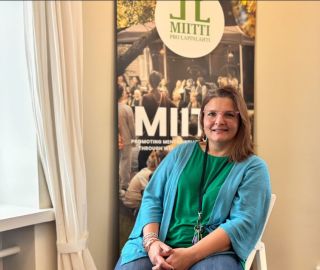 Nainen istuu MIITTI-julisteen edessä tuolissa ja hymyilee