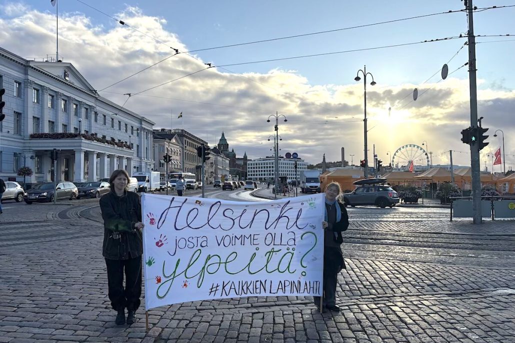 Kaksi ihmistä kannattelee banderollia Helsingin kaupungintalon edustalla auringon noustessa.