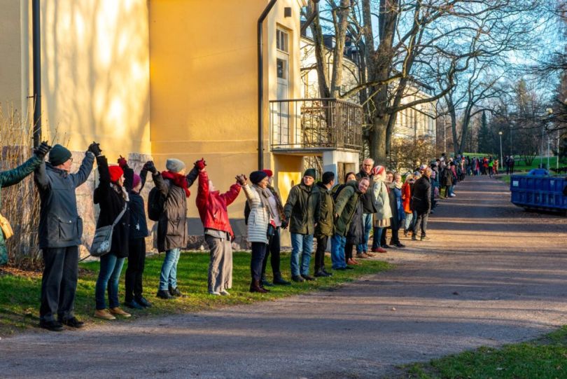 Vuoden 2018 Muuri-tapahtumassa päärakennuksen seinän edessä seisoo ihmisiä käsi kädessä silmänkantamattomiin.