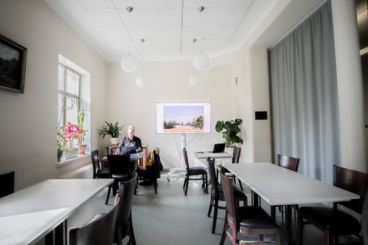 Kuvassa etualalla on pöytiä ja tuoleja. Taaempana on pöydässä istuva henkilö ja Osasto5:n televisio.