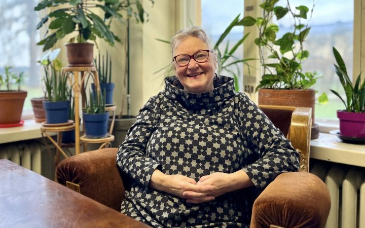 Nainen hymyilee tuolissa viherkasvien ja ikkunan äärellä.