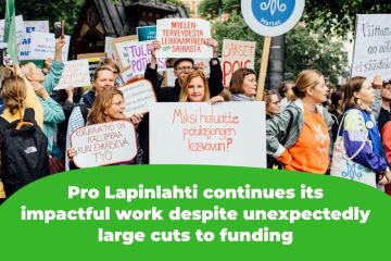 The picture shows protesters holding signs that read, among other things, "Why do you want patient queues to grow?" and "Corrective work is more expensive than preventive work" and "Cutting mental health services is sick."
