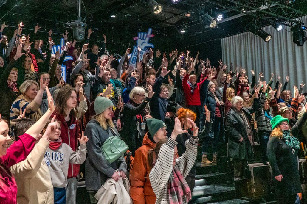 Kuvassa iso sali yleisöä kädet ilmassa kirjasto Oodin salissa.